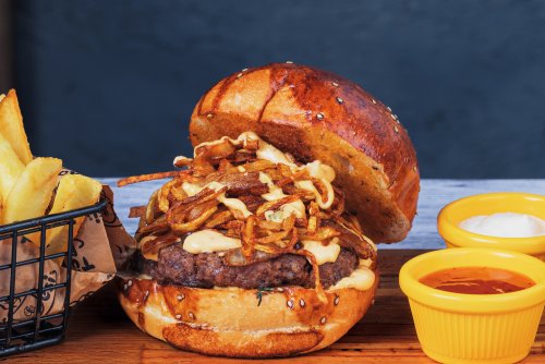 Yiyeceklerin üzerine düşen yumuşak ışık, hamburgerin detaylarını ve dokusunu belirginleştiriyor, özellikle susamlı ekmek, erimiş peynir ve çıtır soğan halkalarının parlaklığını öne çıkarıyor.Renkler açısından, hamburgerin altın rengi tonları, patates kızartmasının sıcak sarılığı ve kırmızı sos kapları ile dengeleniyor. Renklerin bu şekilde kullanımı, tabağın genelinde iştah açıcı ve cezbedici bir görünüm yaratıyor.Kamera açısı ise yemeği hafif yukarıdan ve yakın plandan çekerek, tabağın tamamını net bir şekilde gösteriyor. Bu açı, yemeğin tüm bileşenlerini görsel olarak dengeli bir şekilde sunar ve izleyicinin dikkatini yemeğin merkezine çeker. Fotoğraf, yemeğin hem lezzetli hem de estetik görünmesini sağlıyor.
